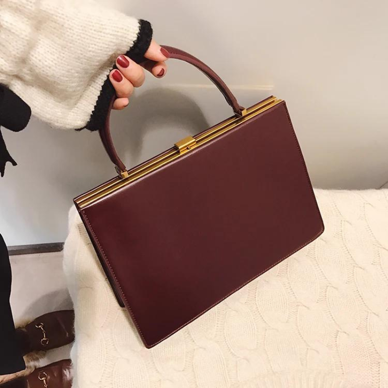 Main aux ongles rouges saisissant l'anse du sac bordeaux, posé sur un plaid crème