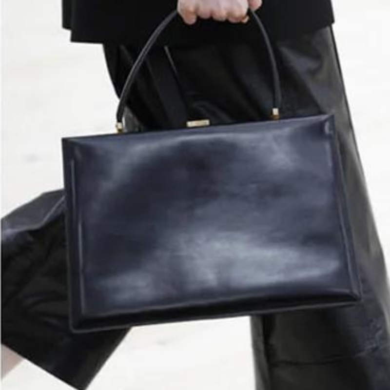 Tenu par l'anse au-dessus d'un pantalon noir, le sac rectangulaire brille d'un noir profond