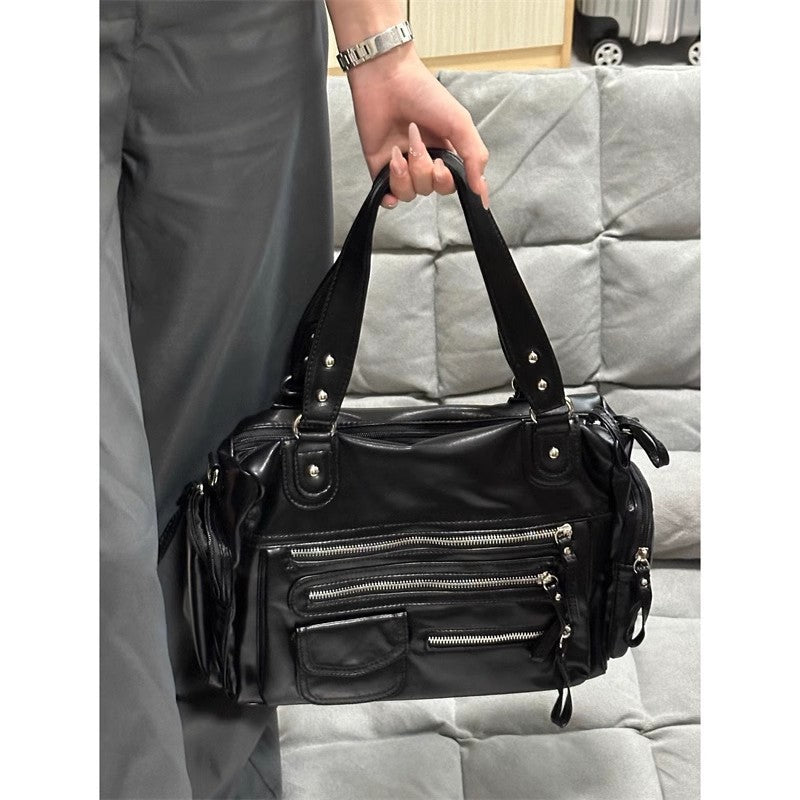 À la main devant un canapé gris, le grand sac noir souple laisse pendre sa bandoulière